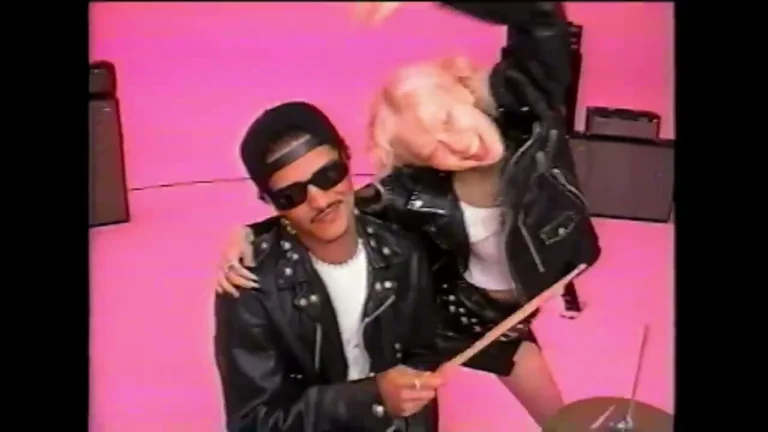 Two performers in black jackets beside a drum kit against a pink studio backdrop