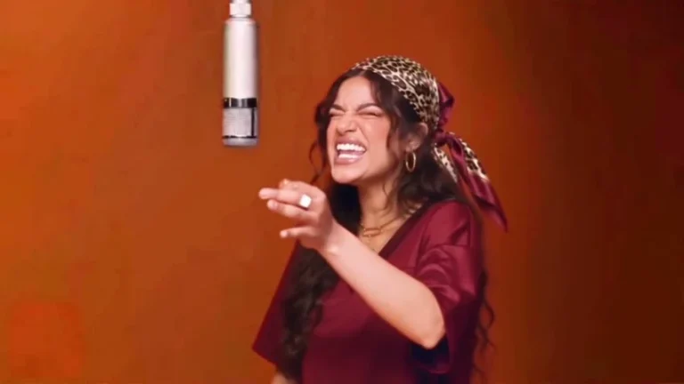 A singer in burgundy gestures under a hanging microphone against an orange studio wall