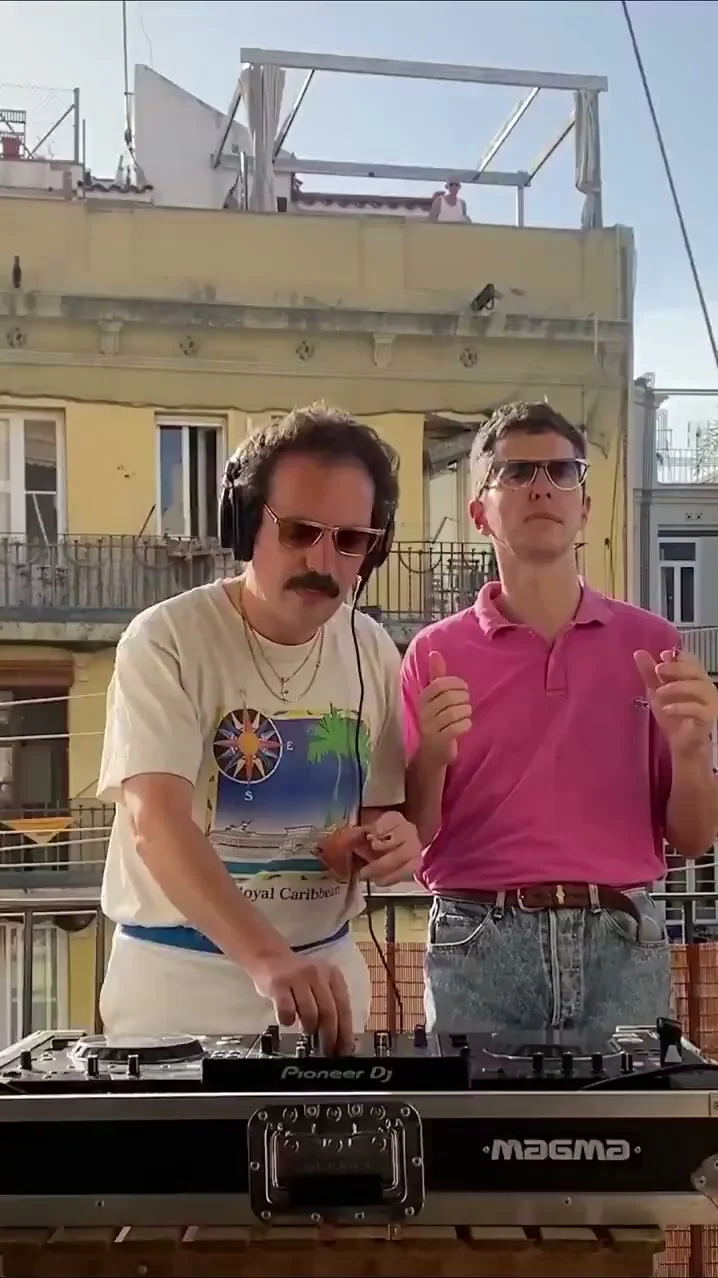 Two performers at DJ decks on a sunny balcony, with a yellow building behind them