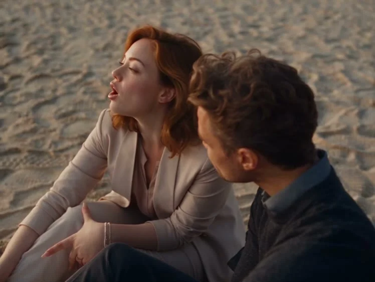 Two people sit together on a sandy beach, one singing while the other looks toward them