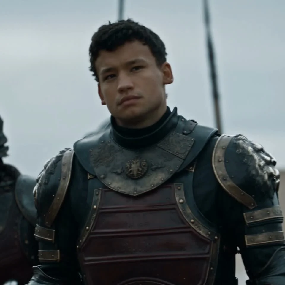 A dark-haired person in dark metal armor facing the camera against a pale sky