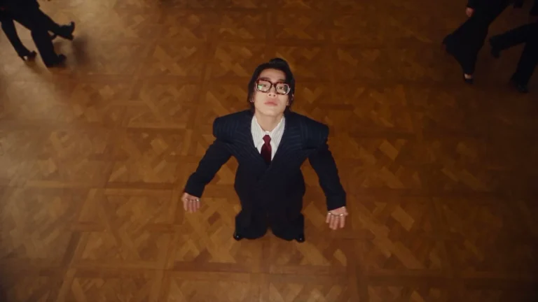 Overhead view of a person in a dark pinstripe suit and glasses on a patterned wooden floor, with other figures at the edges.