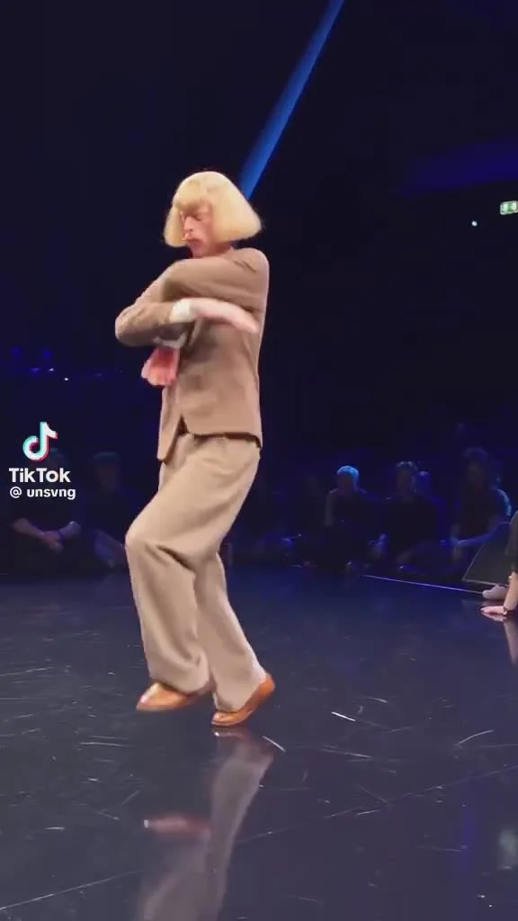 Breaking dancer in a loose suit and blond wig on a dark stage, with the crowd behind