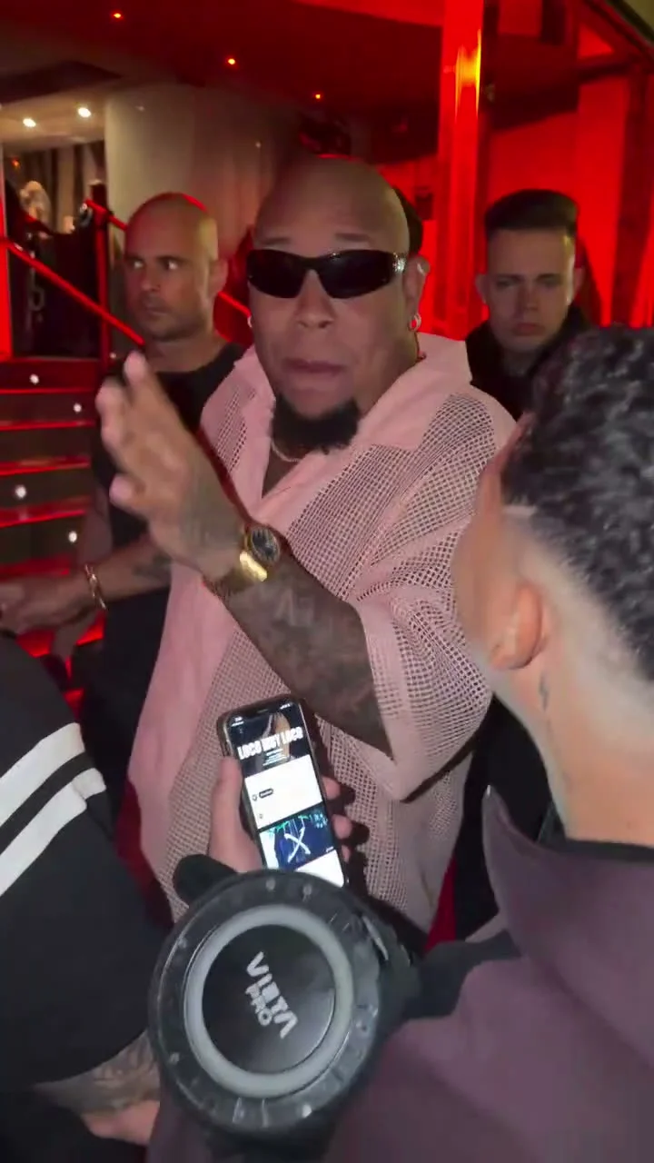 Source preview of a rapper in a pale pink shirt and black sunglasses performing in a red-lit crowd near a staircase