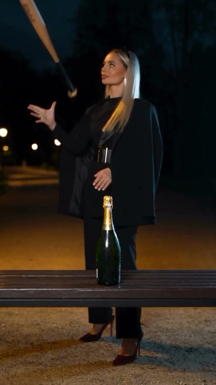 Woman in black at night tossing a bat above a champagne bottle on a wooden bench