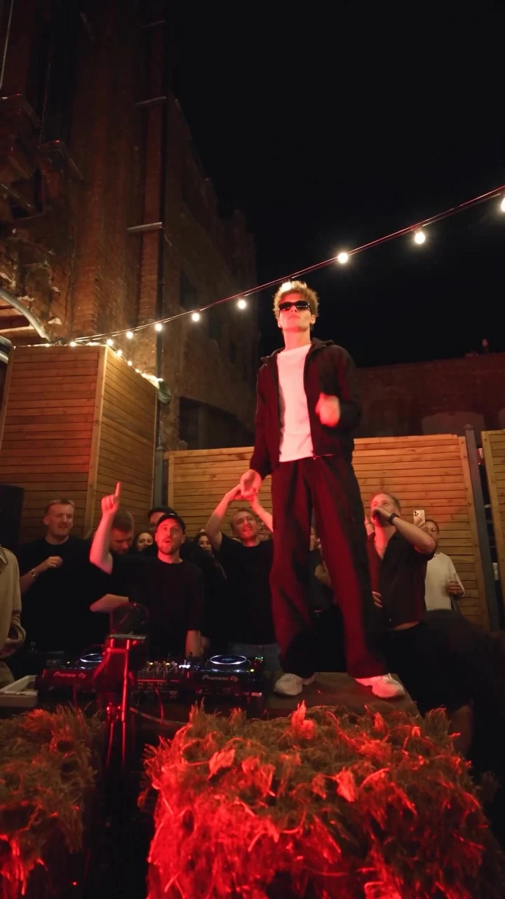 Dancer in sunglasses standing above a DJ booth at a nighttime courtyard party under string lights and red lighting