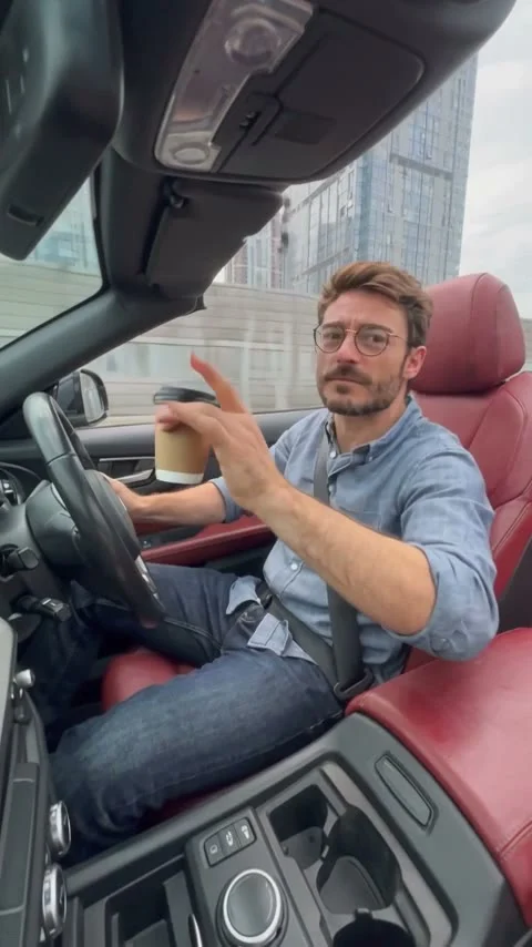 Man in glasses and a blue shirt seated beside red car upholstery, holding a takeaway cup toward the camera