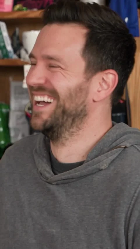 Close-up of a dark-haired man in a gray hoodie laughing beside shelves.