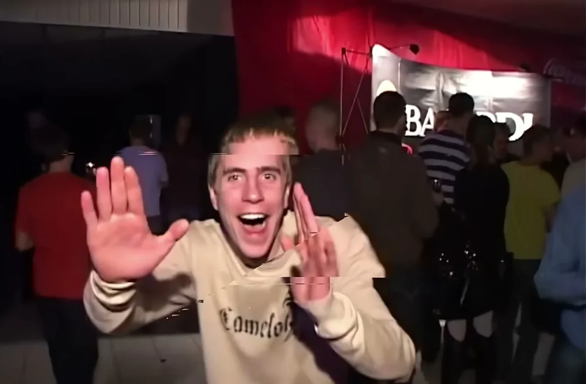 Man in a light sweater raises both hands toward the camera in a crowded red-lit club.