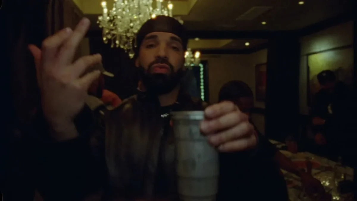 A bearded man holds a silver cup toward the camera in a dim banquet room with a chandelier and guests behind him.