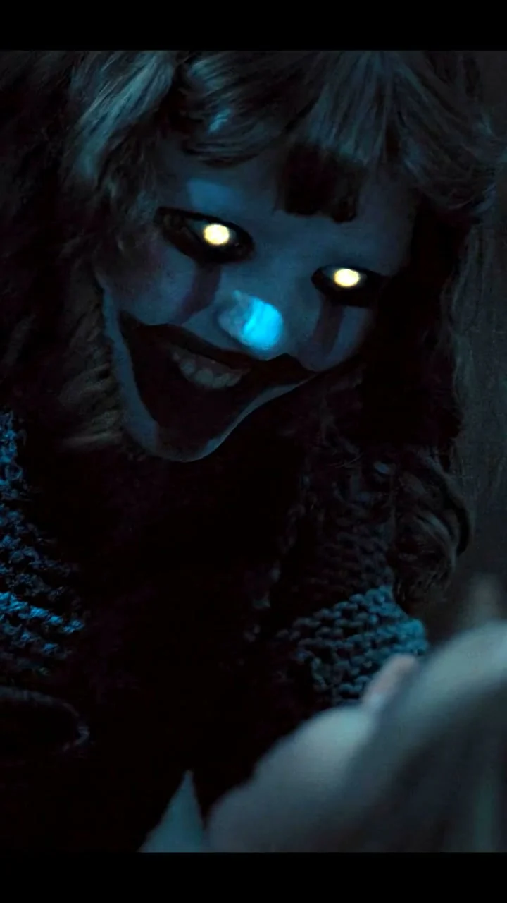 Dark close-up of a masked creature with glowing yellow eyes and a blue-lit nose reaching toward someone