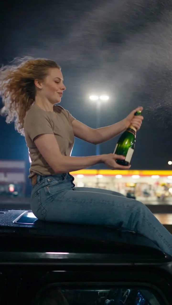 Person with windblown hair holding a green bottle while seated on a dark vehicle roof at night