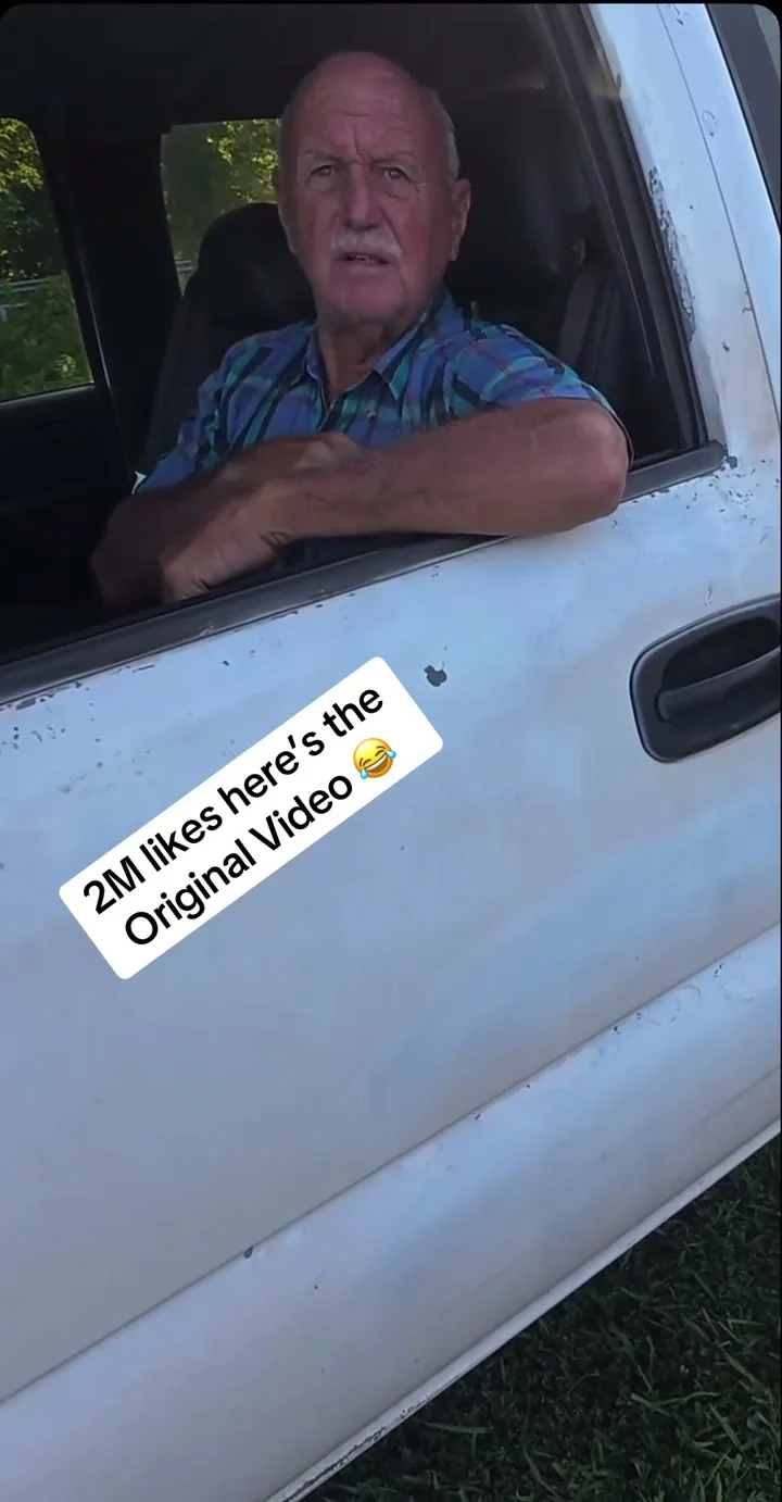 Man in a blue plaid shirt resting his arms on the window of a white pickup, with a tilted white text sticker on the door