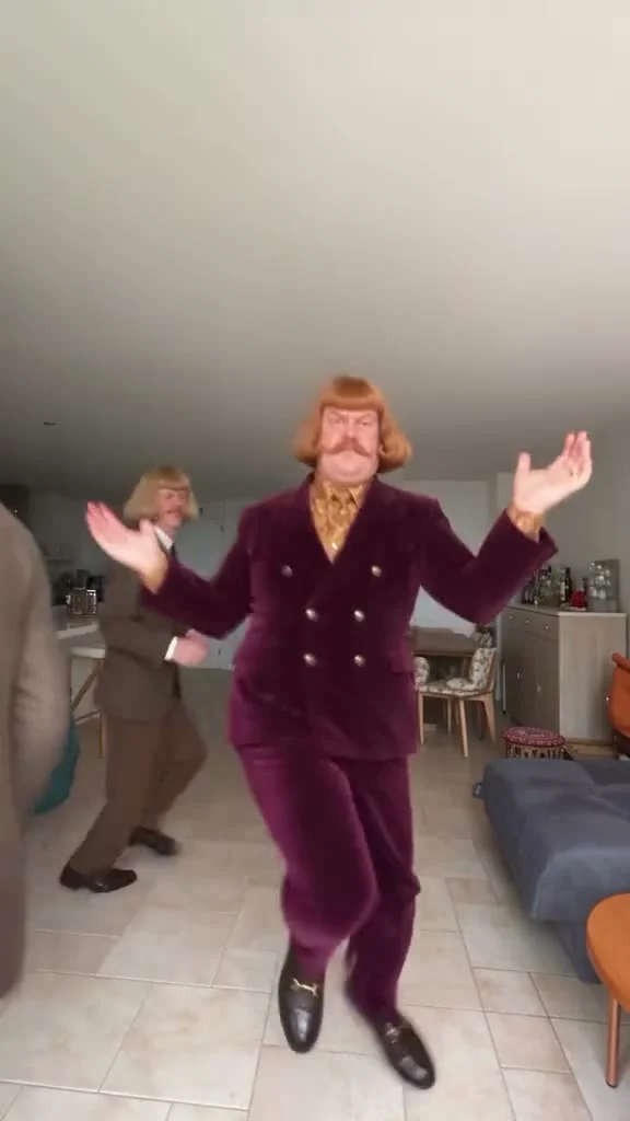 A mustached dancer in a burgundy suit raises his hands near the camera while another suited dancer stands in the background of a furnished room.