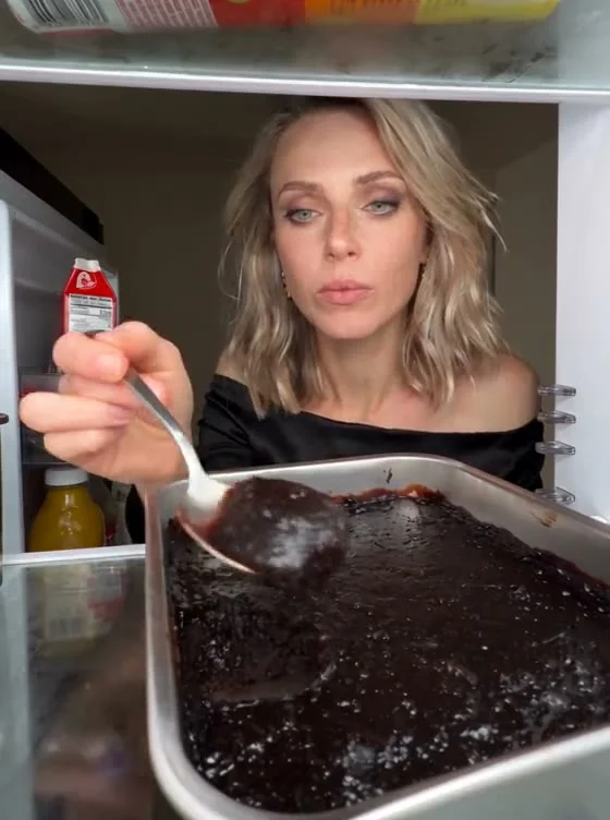 Woman in a black off-shoulder top holding a spoon over a dark chocolate cake tray, viewed from inside a refrigerator