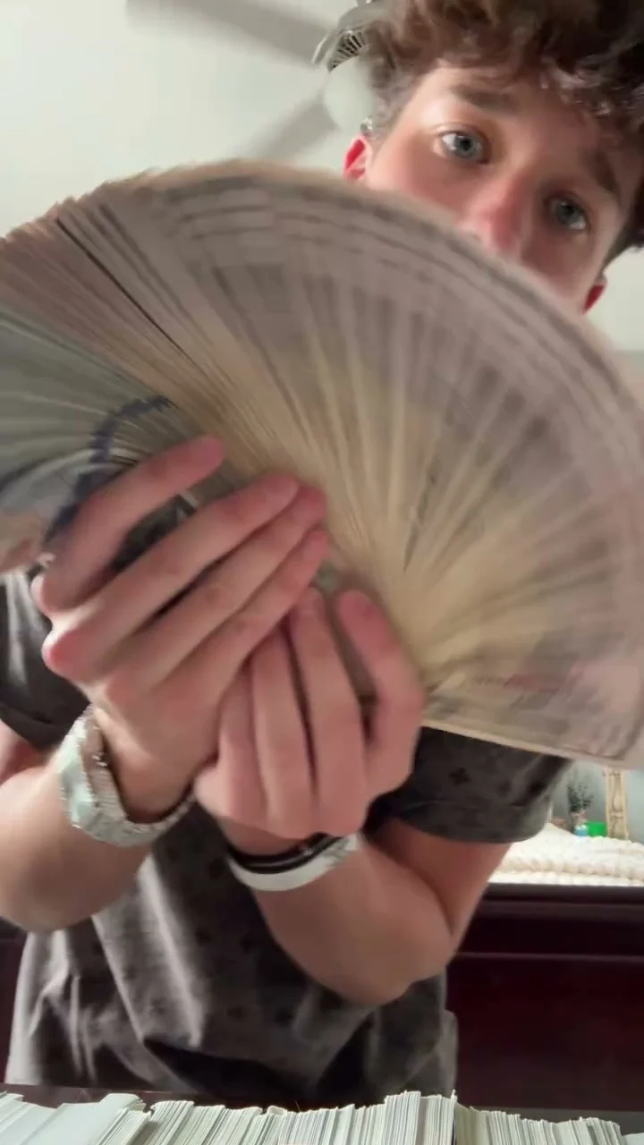 Person holds a wide fan of banknotes toward the camera with stacks of cash visible along the bottom of the frame