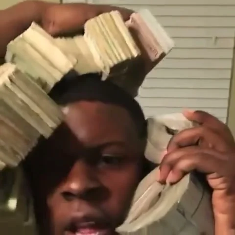 Performer holds a bundled stack of cash to his ear with more cash raised over his head in front of window blinds