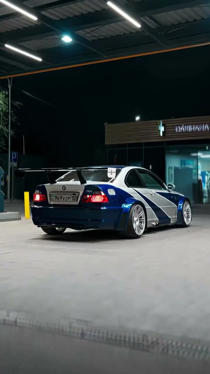 Blue and silver BMW M3 GTR-style car at a lit gas station at night, seen from the rear