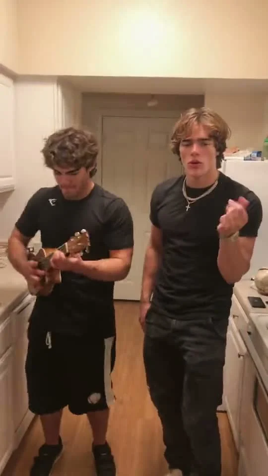 Two people in dark clothes stand in a narrow kitchen; the person on the left holds a small stringed instrument.