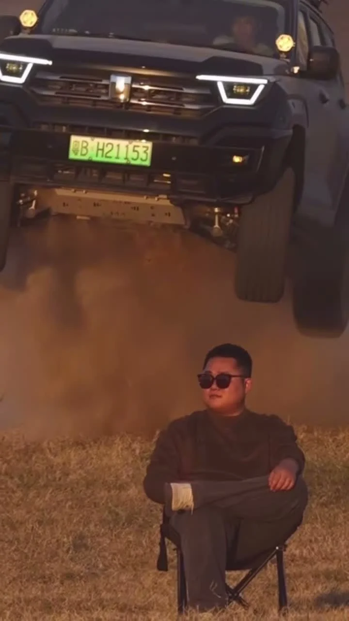 A dark pickup truck airborne above a seated man in sunglasses on dusty ground