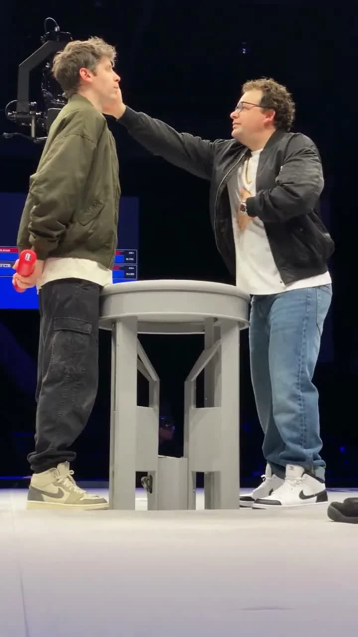Two people facing each other across a stage table as the person on the right reaches toward the person on the left
