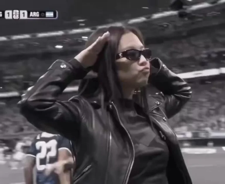 Source preview of a person in dark sunglasses and a black leather jacket posing on a football pitch, with a player and scoreboard behind