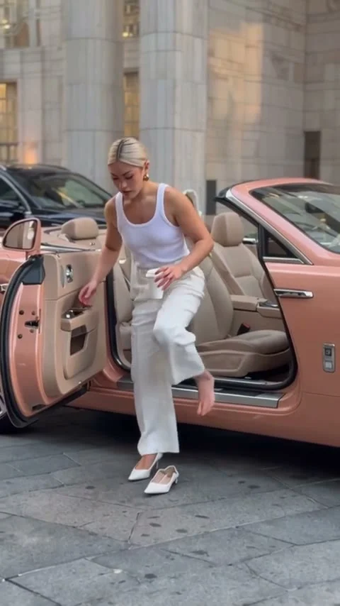 Person in white top and light pants stepping out of a pink convertible beside white shoes on the pavement outside a stone building