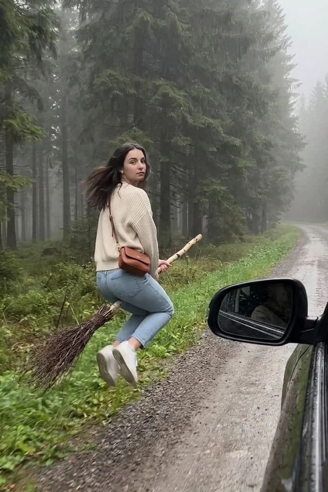Woman in a cream sweater on a twig broom beside a car on a misty forest road